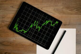 Flatlay of an iPad displaying stock market graph on a wooden desk with a pencil and paper.