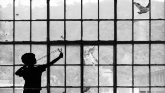 Black and white silhouette of a child aiming a slingshot by a large glass window.